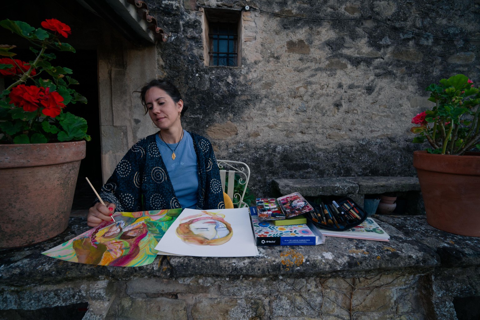 Laia painting with watercolours outdoors in front of a stone wall.